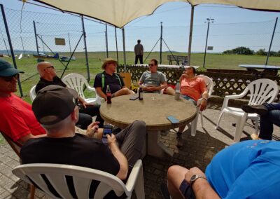 Gruppe von Vereinsmitgliedern und Gästen des MFC Riebelsdorf sitzt entspannt auf weißen Kunststoffstühlen um einen runden Steintisch unter einem Sonnensegel. Im Hintergrund ist der Modellflugplatz mit Start- und Landebahn sowie ein Windrichtungsanzeiger zu sehen. Die Teilnehmenden genießen das sonnige Wetter und führen entspannte Gespräche