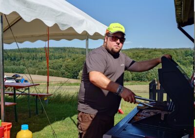 Ein Vereinsmitglied des MFC Riebelsdorf steht am Grill unter freiem Himmel und bereitet Würstchen zu. Er trägt eine gelbe Kappe mit der Aufschrift „Flugleiter“ und eine Sonnenbrille. Im Vordergrund steht eine Biertischgarnitur mit Senf und Grillzubehör. Im Hintergrund sind ein Festzelt, grüne Wiesen und bewaldete Hügel zu sehen.