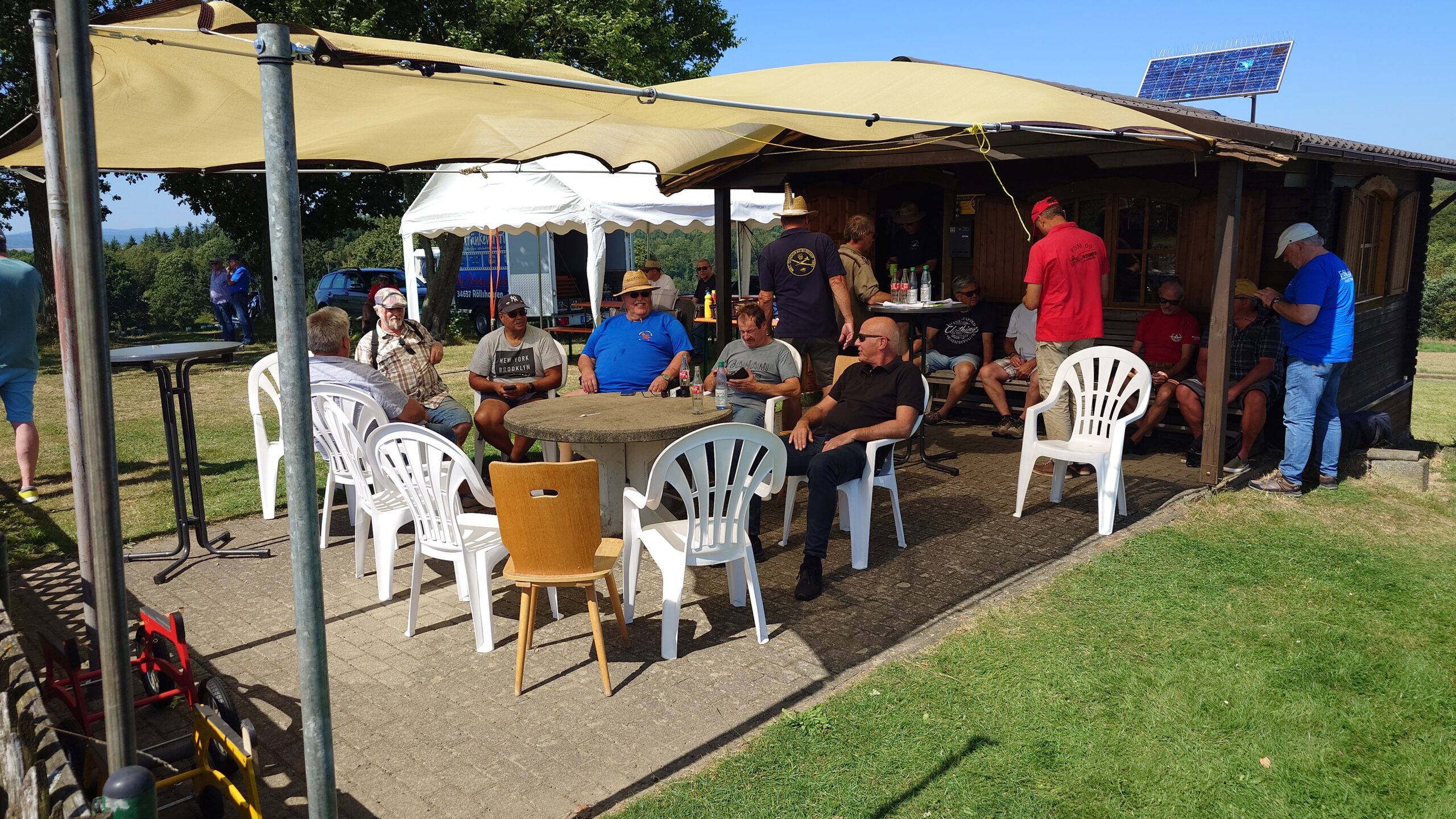 Geselliges Beisammensein beim Sommerfest des MFC Riebelsdorf: Vereinsmitglieder und Gäste sitzen auf weißen Kunststoffstühlen und Holzbänken rund um einen Steintisch vor dem Vereinsheim. Das Vereinsheim ist teilweise überdacht und bietet Schutz vor der Sonne. Über dem Eingang ist eine kleine Solaranlage zu sehen. Im Hintergrund stehen Festzelte und Parkplätze. Die Atmosphäre ist entspannt und fröhlich