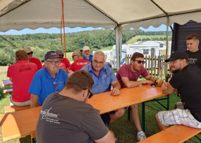 Gruppe von Vereinsmitgliedern und Gästen des MFC Riebelsdorf unter einem Festzelt. Sie sitzen an orangefarbenen Biertischgarnituren, unterhalten sich und genießen das gesellige Beisammensein bei schönem Wetter. Im Hintergrund sind Wohnmobile und die grüne Umgebung des Modellflugplatzes zu sehen